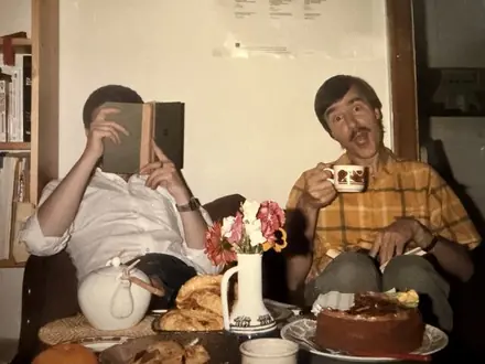 Two friends at a table with tea and cake; one hides behind an open book, Robert grins over a mug.
