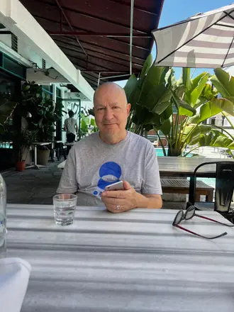 Robert at a patio table under an awning in June 2026, glasses folded beside him, palms behind.