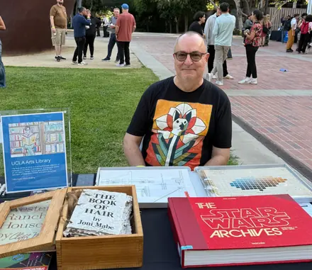 Robert seated behind an outdoor UCLA Arts Library table spread with artists&rsquo; books, including Joni Mabe&rsquo;s The Book of Hair, students crossing the plaza behind him.